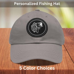 Può includere: Un cappello da baseball grigio con una toppa nera che presenta una silhouette bianca di un pesce con la bocca aperta. La toppa ha un design a corda bianca attorno al bordo. Il testo "Personalized Fishing Hat" è nella parte superiore dell'immagine e "5 Color Choices" nella parte inferiore.