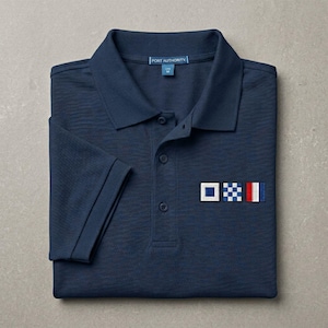 May include: A navy blue polo shirt with a collar, a three-button placket, and short sleeves. The shirt features an embroidered design on the chest consisting of three square nautical signal flags in white, blue, and red. A Port Authority label is visible.