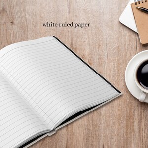 Puede incluir: Un cuaderno blanco con l&iacute;neas y encuadernaci&oacute;n negra abierto sobre una superficie de madera. El cuaderno est&aacute; en blanco y listo para escribir.