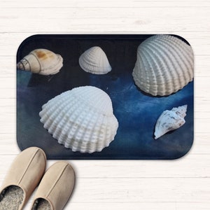 May include: A blue bath mat with five white seashells on it. The shells are arranged in a semi-circle shape. The mat is made of a soft, absorbent material.