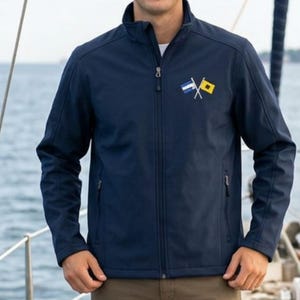 Può includere: Una giacca blu navy con cerniera, collo alto e due tasche laterali con chiusura a zip. Sul petto sinistro è presente un ricamo raffigurante due bandiere nautiche incrociate, una blu e bianca e l'altra gialla e nera.