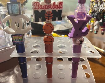 Può includere: Tre penne decorative con diversi ciondoli in cima. I ciondoli sono un logo Chanel bianco, una palla da basket con le parole "Basketball Mom" e un logo LV viola. Le penne sono in un portapenne di plastica trasparente.