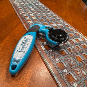 Pode incluir: Um cortador rotativo azul e preto com o texto "TrueCut" e uma régua de quilting transparente com linhas de grade. O cortador é projetado para um corte fácil, seguro e preciso. A régua tem medidas em centímetros.