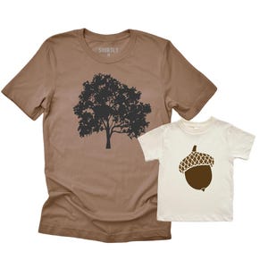 May include: Two t-shirts, one adult and one child size. The adult shirt is brown with a black tree graphic. The child's shirt is off-white with a brown acorn graphic. The adult shirt has the word "SHIRTLY" printed on the tag.