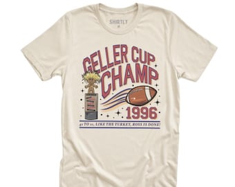 May include: Cream-colored t-shirt featuring the text "GELLER CUP CHAMP 1996" in red and blue. A cartoon football and trophy are also displayed. The shirt also has the text "43 TO 11, LIKE THE TURKEY, ROSS IS DONE!"