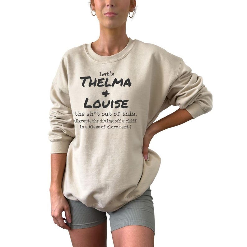 Może przedstawiać: Beżowa bluza z okrągłym dekoltem z czarnym napisem "Let's THELMA + LOUISE the sh*t out of this." Bluza zawiera r&oacute;wnież tekst "(Except, the diving off a cliff in a blaze of glory part.)".