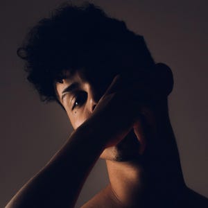 May include: A dramatic, artistic portrait of a person, with the face partially obscured by an arm. The lighting creates strong shadows and highlights, emphasizing the subject's form and features. The image has a moody, introspective feel.