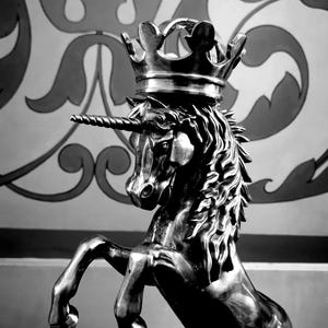 Op de afbeelding: Een zwart-wit sculptuur van een steigerende eenhoorn met een kroon. De eenhoorn heeft een spiraalvormige hoorn en een vloeiende manen. De achtergrond heeft een decoratief patroon in grijstinten.