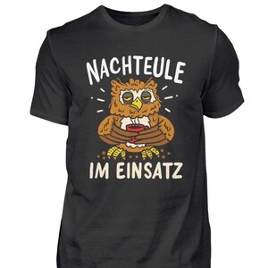 Könnte beinhalten: Schwarzes T-Shirt mit einer Cartoon-Eule, die eine Kaffeetasse hält, und dem Text "NACHTEULE IM EINSATZ" in Weiß. Die Eule ist braun und gelb mit verschlafenen Augen. Das Shirt ist aus weichem Material.