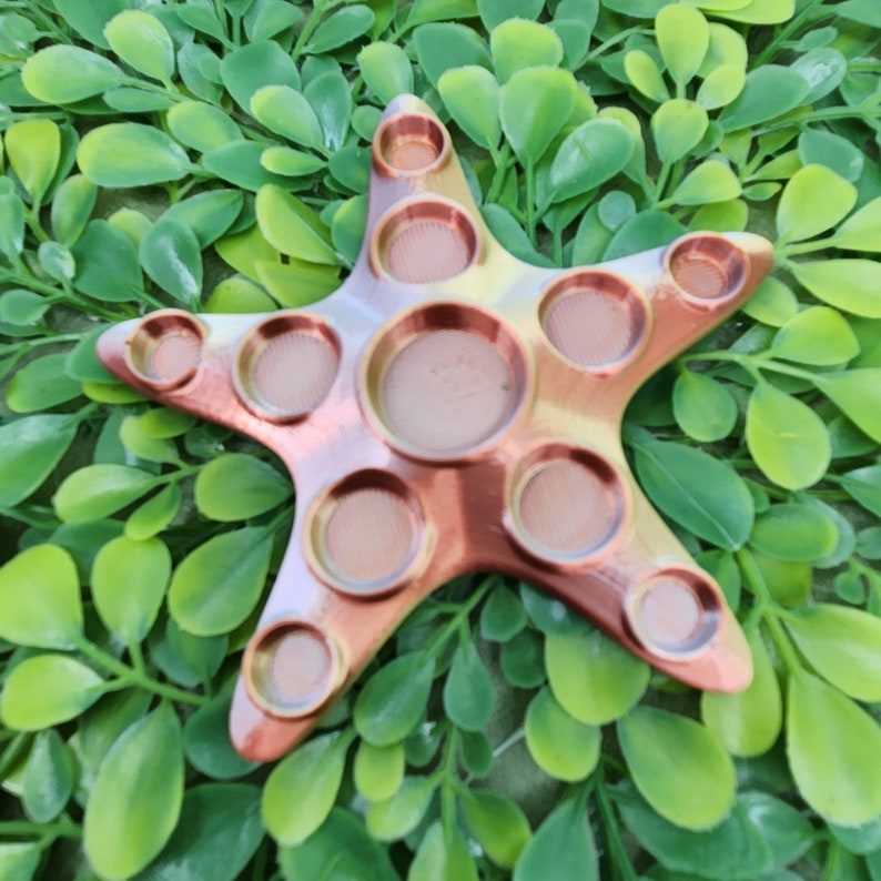 May include: A copper-colored starfish-shaped fidget toy with nine dimpled circles. The toy is made of a smooth, shiny material and has a slightly textured surface.