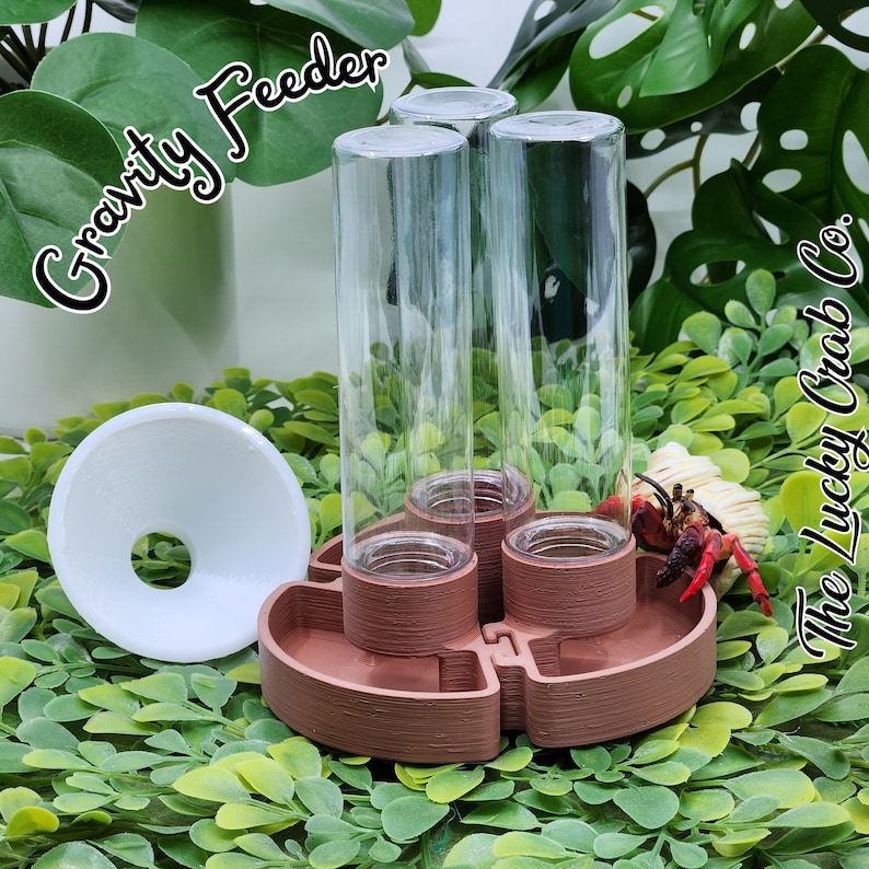 May include: A gravity feeder with three clear glass cylindrical tubes attached to a brown, circular, multi-compartment base. A white plastic funnel sits to the left. The text Gravity Feeder and The Lucky Crab Co. appear in the image.