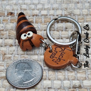 May include: A brown leather keychain with a small, detailed, brown and black leather hermit crab charm. The keychain also has a round brown leather tag with the words "Lucky Crab" engraved on it. The keychain is attached to a silver metal ring and clasp.