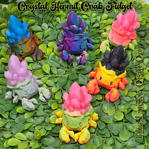 May include: A set of six colorful crystal hermit crab fidget toys. Each crab is made of a different color of resin and has a crystal-like shell. The crabs are all sitting on a bed of green leaves. The text "The Lucky Crab Co." is visible in the image.