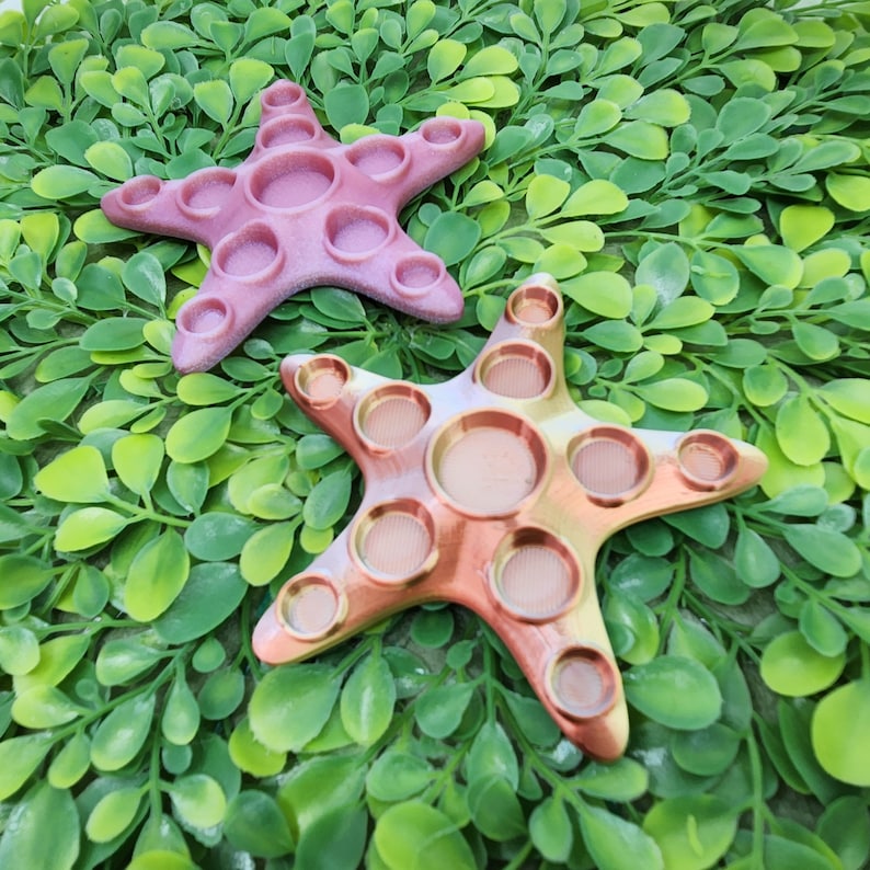 May include: Two colorful silicone starfish-shaped fidget toys. The top toy is pink and the bottom toy is gold. Both toys have multiple dimples that can be pushed in and out.
