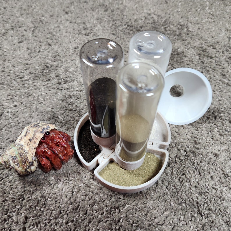 May include: A beige circular feeding station with three clear vertical cylinders containing different colored sands, accompanied by a small hermit crab with a textured tan shell and a separate white plastic funnel accessory.