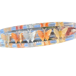 Op de afbeelding: Transparante ovale haarclip met zes vlinderaccenten in oranje, wit en geel. De clip heeft een rechthoekig ontwerp met kleurrijke accenten. Een stijlvol accessoire voor elk kapsel.