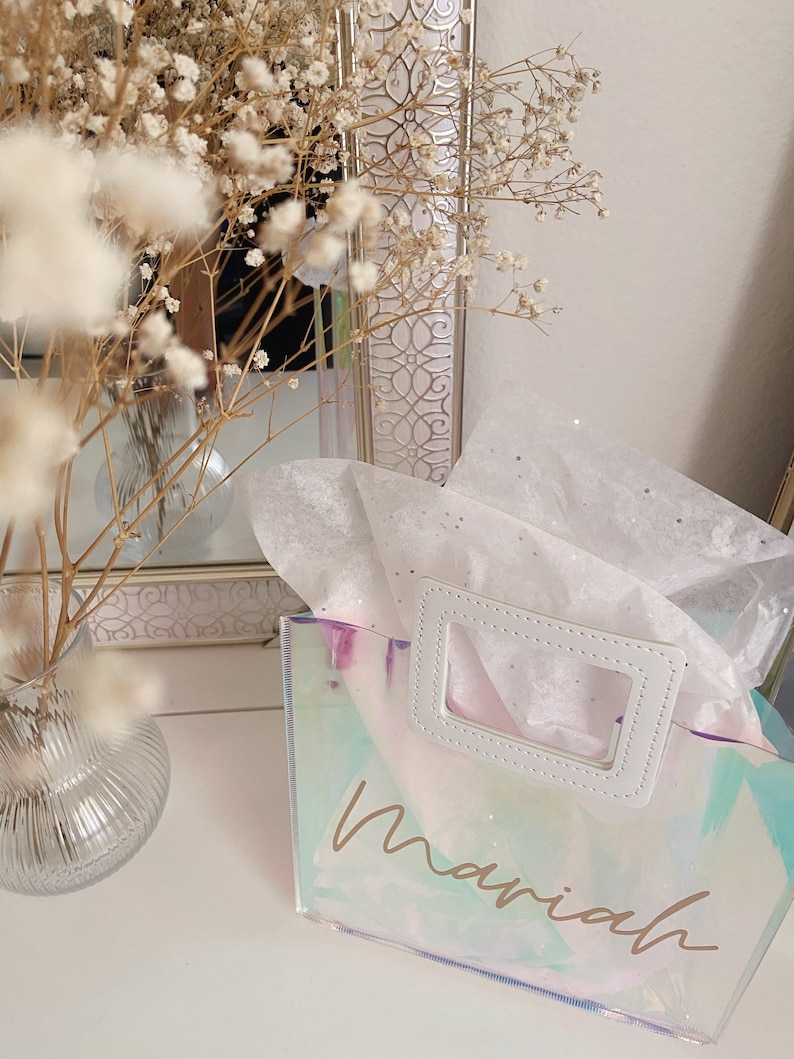 May include: A clear iridescent tote bag with a white handle and the name "Mariah" written in gold script.