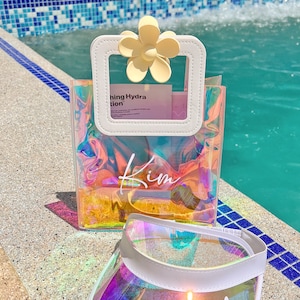 May include: A clear iridescent tote bag with a white flower-shaped handle and the word "Kim" written on it. A clear iridescent visor with a white brim and the word "Bride" written on it.