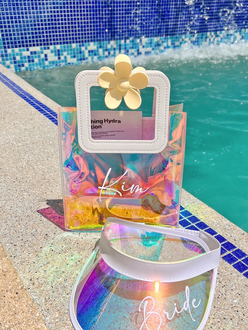 May include: A clear iridescent tote bag with a white flower-shaped handle and the word "Kim" written on it. A clear iridescent visor with a white brim and the word "Bride" written on it.
