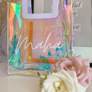 May include: A clear iridescent tote bag with a white leather handle and the name "Maha" written in white script. The bag is sitting on a white surface with white and pink roses.