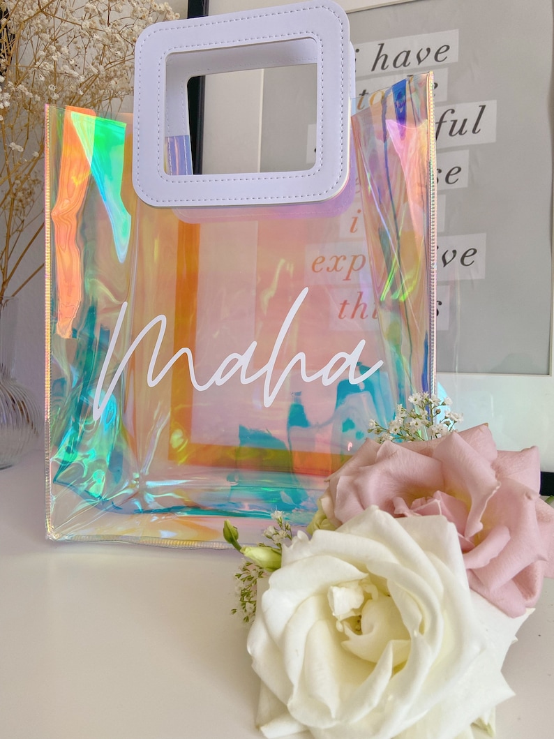 May include: A clear iridescent tote bag with a white leather handle and the name "Maha" written in white script. The bag is sitting on a white surface with white and pink roses.
