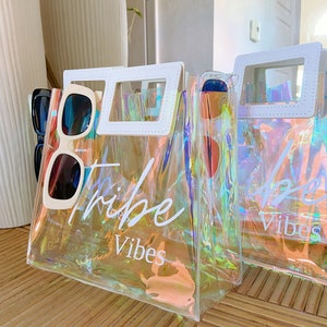 May include: Two iridescent clear plastic tote bags with white handles and white text. The bag on the left says "Tribe Vibes" and the bag on the right says "Good Vibes".
