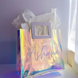 May include: A clear iridescent gift bag with a purple handle and a white inscription that says "Ashara".