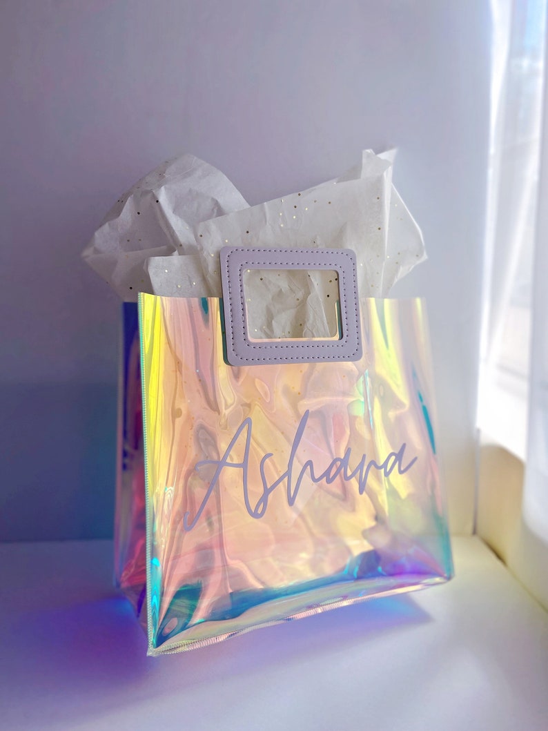 May include: A clear iridescent gift bag with a purple handle and a white inscription that says "Ashara".