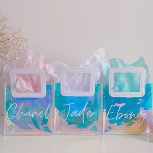 May include: Three iridescent clear plastic gift bags with white handles and white lettering. The bags are filled with tissue paper and have the names "Chanel", "Jade", and "Ebony" written on them.