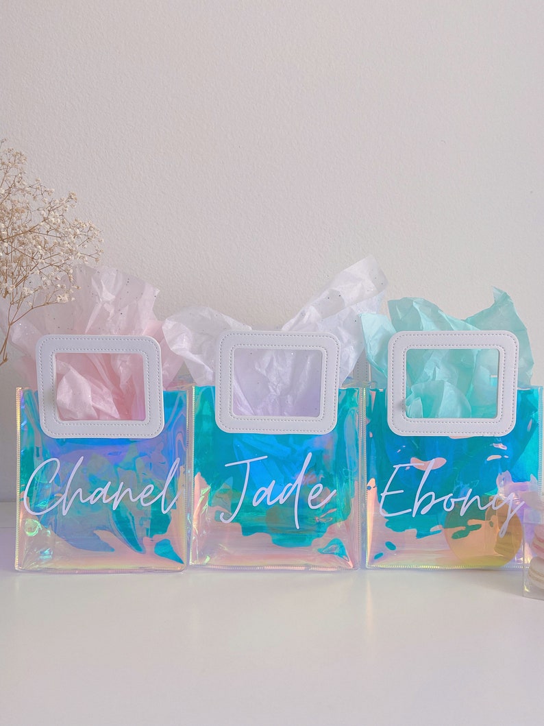 May include: Three iridescent clear plastic gift bags with white handles and white lettering. The bags are filled with tissue paper and have the names "Chanel", "Jade", and "Ebony" written on them.