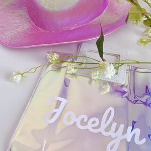 May include: A clear iridescent tote bag with a white handle and the name "Jocelyn" written in white cursive letters. The bag is sitting on a white surface with a pink iridescent cowboy hat and white flowers.
