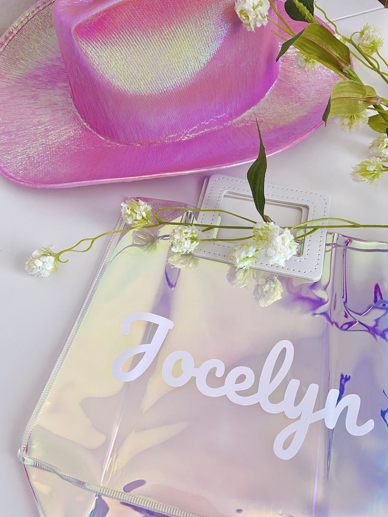 May include: A clear iridescent tote bag with a white handle and the name "Jocelyn" written in white cursive letters. The bag is sitting on a white surface with a pink iridescent cowboy hat and white flowers.