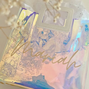 May include: A clear iridescent tote bag with a white leather handle and a clear window. The bag has the name "mariah" written in gold lettering.