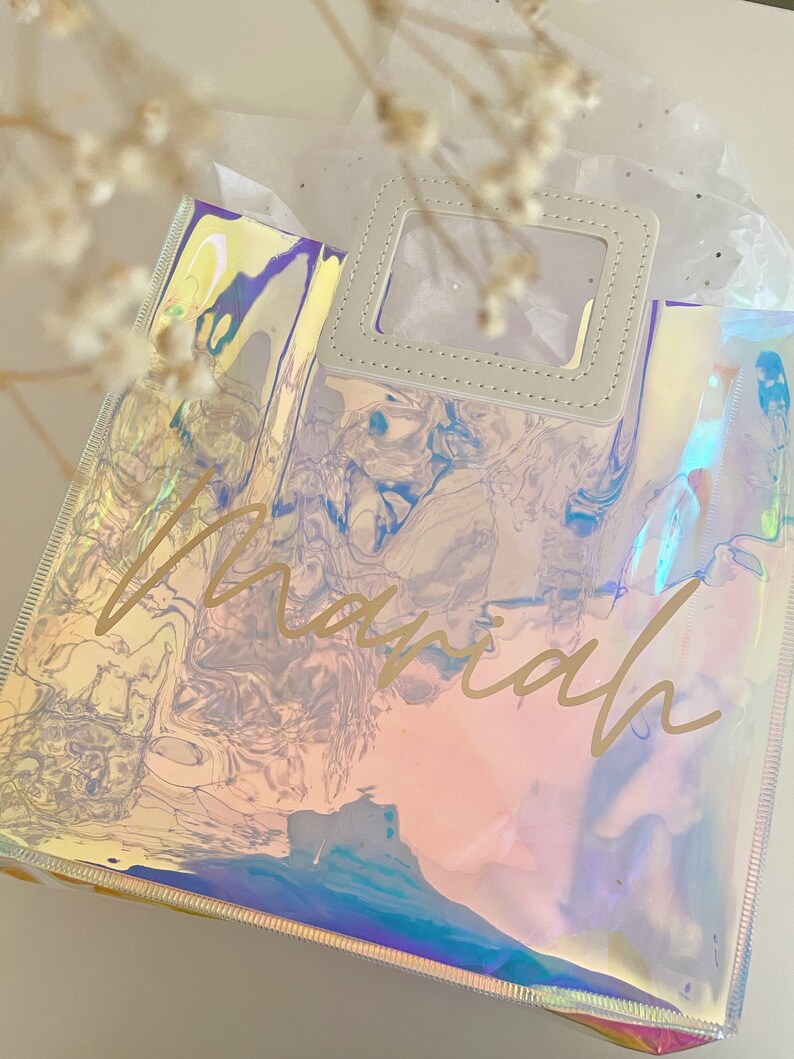 May include: A clear iridescent tote bag with a white leather handle and a clear window. The bag has the name "mariah" written in gold lettering.