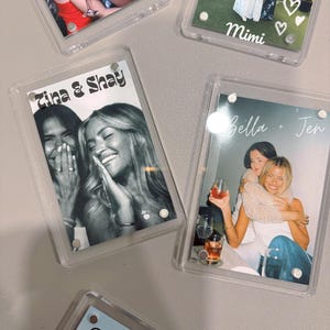 Peut inclure: Quatre cadres en plastique transparent avec des photos et du texte à l'intérieur. Les cadres sont accrochés à un mur. Les photos représentent deux femmes dans chaque cadre. Le texte dans les cadres indique "Tina & Shay", "Bella + Jen", "Mimi" et un cœur.