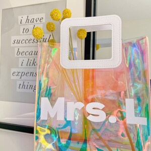 May include: A clear iridescent tote bag with a white handle and the text "Mrs. L" printed on the front.