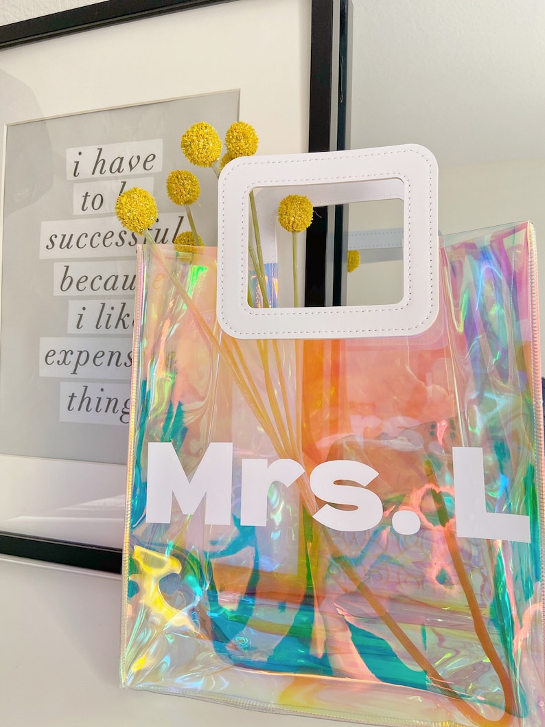 May include: A clear iridescent tote bag with a white handle and the text "Mrs. L" printed on the front.