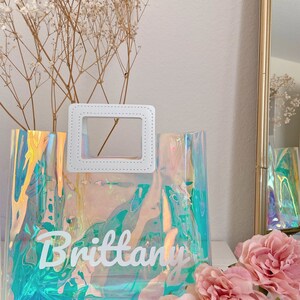 May include: A clear iridescent tote bag with a white handle and the name "Brittany" written in white on the front.
