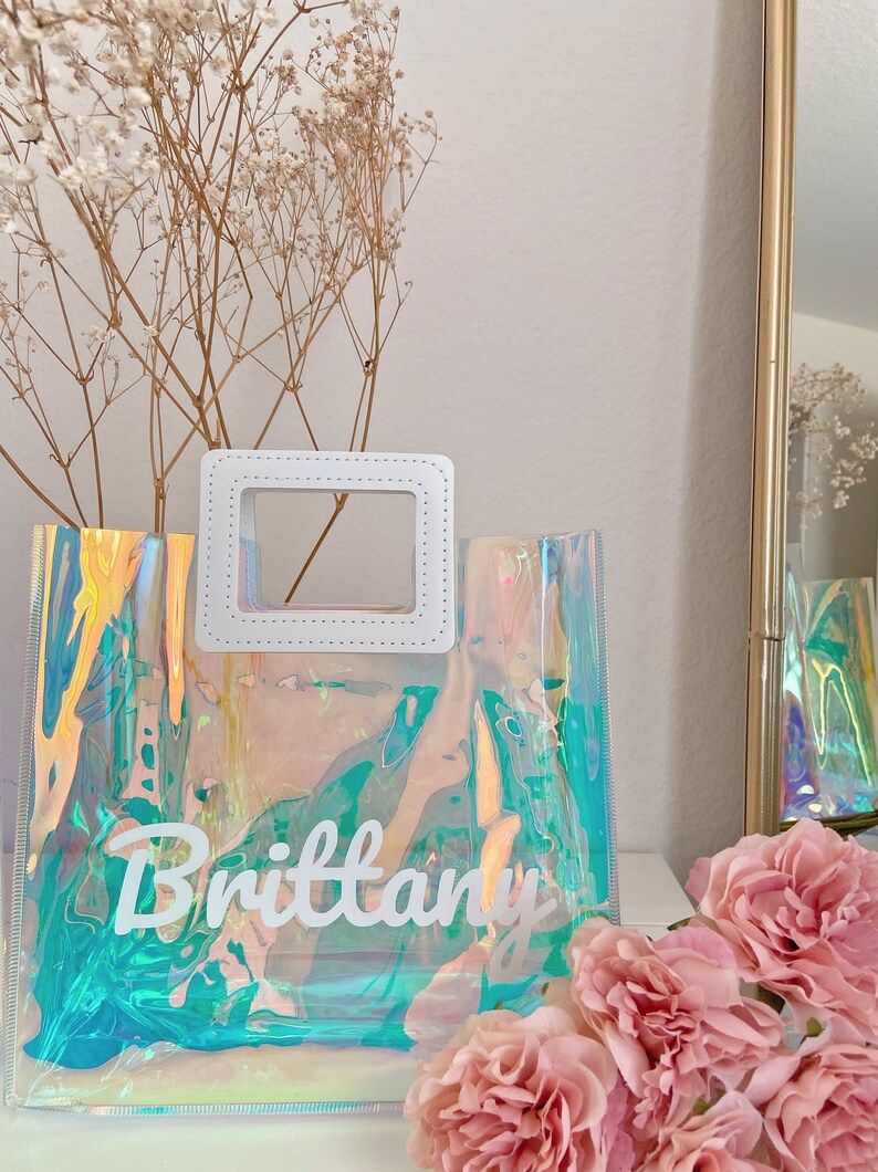 May include: A clear iridescent tote bag with a white handle and the name "Brittany" written in white on the front.