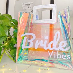 May include: A clear iridescent tote bag with a white handle and the text "Bride Vibes" printed on the front.