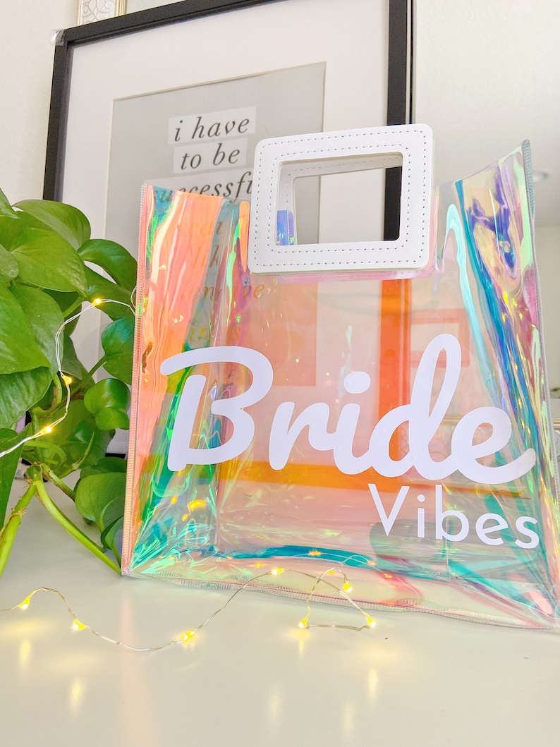 May include: A clear iridescent tote bag with a white handle and the text "Bride Vibes" printed on the front.