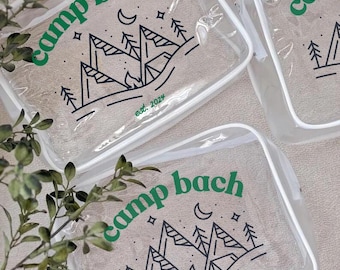 May include: Three clear plastic zippered pouches with white trim. Each pouch features a black and white graphic of mountains, a campfire, and a crescent moon. The text "camp bach est. 2024" is printed in green on two of the pouches.