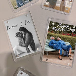 May include: Four clear acrylic photo frames with different images and text. The frames are on a white surface. The text in the frames includes "We love you Mom", "Mama & Me", "Happy Mother's Day", and a blank frame.