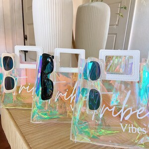 May include: Three iridescent clear plastic tote bags with white handles and white stitching. Each bag has a pair of sunglasses inside and the text "Tribe Vibes" printed on the front.