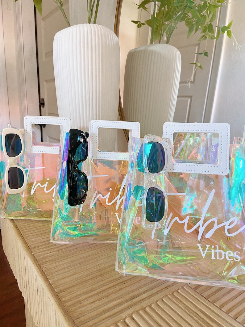 May include: Three iridescent clear plastic tote bags with white handles and white stitching. Each bag has a pair of sunglasses inside and the text "Tribe Vibes" printed on the front.