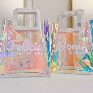 May include: Two iridescent clear plastic tote bags with white leather handles and white text. The bag on the left says "Felicia Maid of Honor" and the bag on the right says "Joni Bridesmaid".