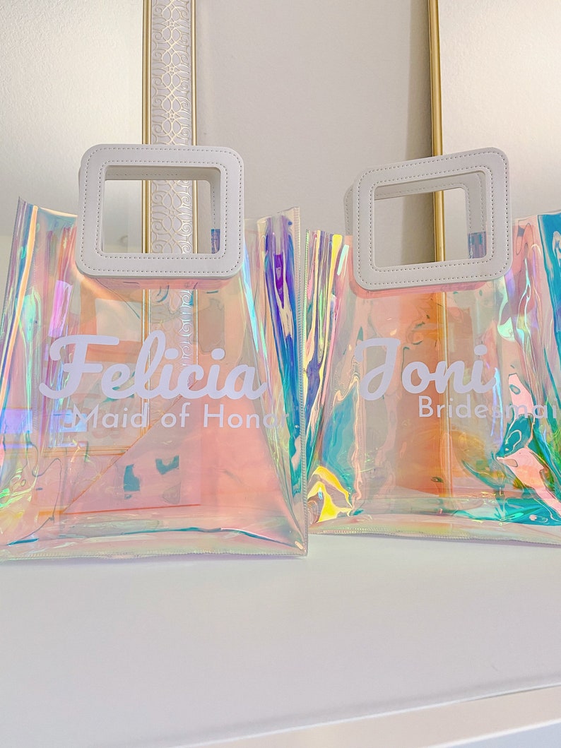 May include: Two iridescent clear plastic tote bags with white leather handles and white text. The bag on the left says "Felicia Maid of Honor" and the bag on the right says "Joni Bridesmaid".
