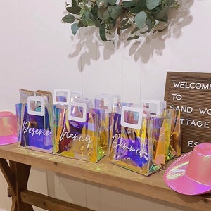 May include: Five iridescent clear plastic bags with white handles and white lettering. The bags are labeled "Desiree", "Nancy", "Summer", "Ashley", and "Sarah". The bags are sitting on a wooden table with a pink cowboy hat and a wooden sign that says "Welcome to Sand Wo Cottage Estates".