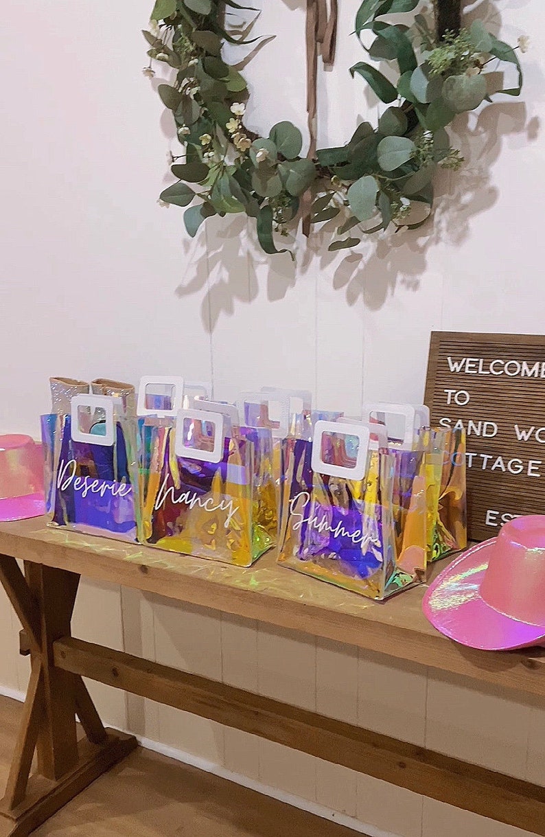 May include: Five iridescent clear plastic bags with white handles and white lettering. The bags are labeled "Desiree", "Nancy", "Summer", "Ashley", and "Sarah". The bags are sitting on a wooden table with a pink cowboy hat and a wooden sign that says "Welcome to Sand Wo Cottage Estates".