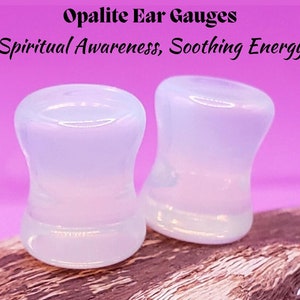 May include: Two white opalite ear gauges, also known as plugs, are shown on a wooden surface. The gauges are smooth and have a slightly iridescent sheen. The text "Opalite Ear Gauges" and "Spiritual Awareness, Soothing Energy" is visible above the gauges.
